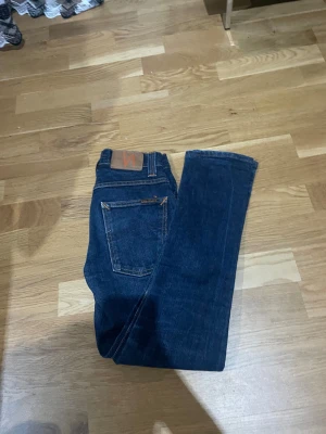 Mörkblå jeans från Nudie Jeans, 27/32 - Snygga mörkblå jeans från Nudie Jeans i klassisk straight fit. Jeansen har orange kontrastsömmar, fem fickor och läderpatch med Nudie-logga bak. Tillverkade i slitstarkt bomullsjeansmaterial, perfekta för en clean och tidlös look.