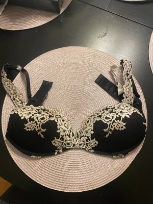 Svart intimissimi bh - Säljer en svart intimissimi bh med beige blommig spetsdetalj. Axelbanden är dekorerade med samma spets och kan justeras. Kuporna är push up och ger snygg passform!🌸Säljer dennq eftersom den är fel storlek för mig!💞