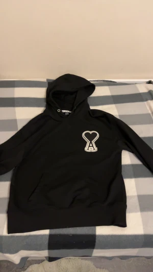 Svart hoodie från AMI med hjärtdetalj - Svart hoodie från AMI med stor vit broderad hjärtdetalj och bokstaven A på bröstet. Tröjan har huva med snörning och klassisk känguruficka framtill. Perfekt för dig som gillar stilrena streetwear-plagg med unik design.