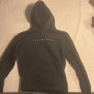 Svart hoodie från Jack & Jones - Svart hoodie från Jack & Jones med vit logotyp på bröstet. Klassisk design med huva och stor magficka framtill. Mjukt material som känns skönt mot huden och långa ärmar med ribbade muddar. Perfekt för chill dagar eller när du vill ha en enkel stil.