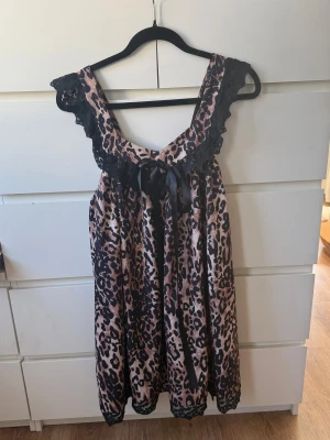 Leopardmönstrad kortklänning med spets - Säljer en leopardmönstrad kortklänning med svarta spetsdetaljer vid axelband och nederkant. Klänningen har en loose passform och ärmlös design, samt en svart rosett framtill. 
