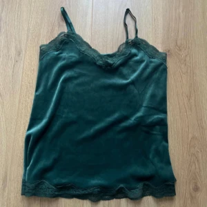 Sammets linne i mörkgrönt med spets - Snyggt mörkgrönt sammets linne med spets från Hunkemöller. Toppen har tunna axelband och vacker spetsdetalj längs halsringning och nederkant. Materialet är mjukt och har en lyxig känsla, perfekt för att lyfta din outfit. Aldrig användt