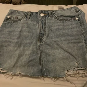 Ljusblå jeanskjol med fransig kant - Säljer en ljusblå jeanskjol med fransig och sliten kant längst ner. Kjolen har klassiska fem fickor, bälteshällor och knappstängning framtill. Baksidan har två fickor med slitna detaljer för en cool, avslappnad vibe.