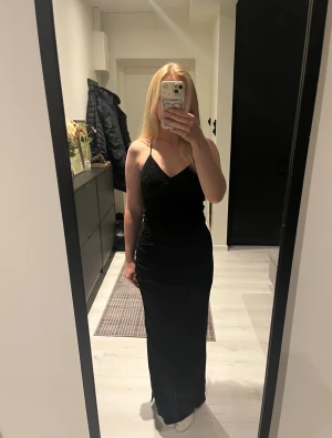 Svart glittrig långklänning med slits - Säljer en svart glittrig långklänning med smala axelband och v-ringning. Ryggen är öppen med fint korsade band. Klänningen har en snygg slits framtill och figurnära passform. Materialet glittrar jättefint, perfekt till fest eller nyårsfirande! 