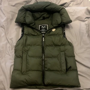 Versace väst  - Snygg mörkgrön dunväst från Versace med hög krage och quiltad design. Västen har dragkedja framtill, två fickor med dragkedja och svarta detaljer längs ärmhålen. Tillverkad i 100% polyester, perfekt för kyliga dagar.