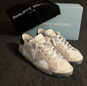 Philippe Model vita sneakers med svart häl - Snygga vita sneakers från Philippe Model med svart häl och beige detaljer. Skorna har snörning med logotyp på skosnörena, rund tå och platt sula. Materialmix av skinn och mocka, samt en silverfärgad logopatch på sidan. Perfekt för dig som gillar exklusiv streetstyle.