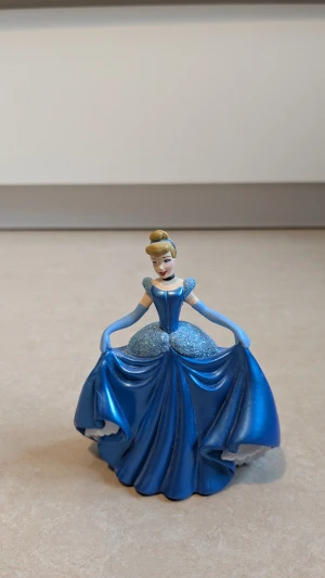  Askungen med blå balklänning med puffärmar - Säljer Askungen med ikonisk blå balklänning. Hela figuren och balklänningen är av plast  Askungen från Disney. Klänningen har glittrigt liv och kjol, puffärmar och markerad midja. Lång modell med vackra draperingar och axelbandslös look. Perfekt för dig som älskar sagofigurer.