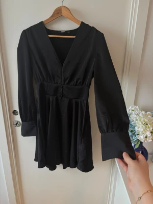 Svart v-ringad klänning Bik Bok  - Super fin svart kortklänning från Bik Bok med v-ringad hals och långa ärmar.   Klänningen har markerad midja, dekorativa knappar framtill och på manschetterna.   Materialet är slätt och har ett elegant fall. Sömnen har gått upp lite fram till, samt knappar som gått bort. Därav är priset billigt om man vill vara den som syr upp och ger klänningen ett nytt liv!   Strl S, men passar även en liten M