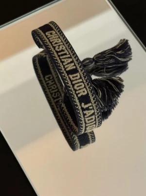 Christian Dior J'adior tyg armband blå/beige - Snyggt armband från Christian Dior med texten 'J'adior' broderad i beige på marinblå bakgrund. Armbandet har justerbar knytning med tofsar i ändarna och är gjort i ett mjukt textilmaterial. Perfekt accessoar för att lyfta din outfit.