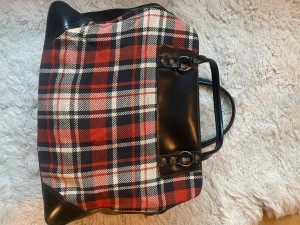 Rutig vintage  handväska med svarta skinnhandtag - Stor och rymlig handväska med rutigt mönster i svart, rött och beige. Väskan har svarta detaljer och handtag i skinn, samt metallnitar vid fästena. Perfekt för dig som gillar klassisk stil med en twist. Materialet är en mix av tyg och skinn. Mått h 35 * 47 cm djup 16 cm.