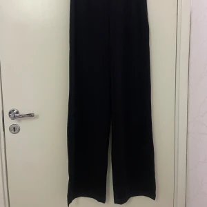 Svarta wide byxor med hög midja - Snygga svarta byxor med hög midja och breda ben. De har en slät linning utan synliga detaljer och är tillverkade i ett mjukt, lite glansigt tyg som ger en stilren look. Perfekta för dig som gillar minimalistisk och trendig stil. Går att klä upp med även använda vardagligt.