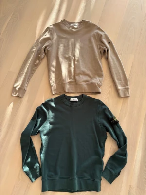 Grön och beige Stone Island sweatshirt storlek S - Mörkgrön och beige sweatshirt från Stone Island med klassisk rund hals och långärmad passform. Tröjan har den ikoniska Stone Island-patchen på vänster ärm och är tillverkad i mjuk bomull. Perfekt för dig som gillar streetwear och vill ha en clean look. Det ena märket / etiketten saknas. Bomull. Fin kvalitet. Båda säljes för 1500:-