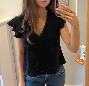 Svart blus med volangärmar och v-ringning - Säljer en svart blus med v-ringad hals och fina volangärmar. Blusen har broderade detaljer och små hålmönster som ger en cool touch. Modellen är lite croppad och har en feminin siluett som passar till jeans eller kjol.
