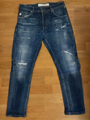 Blå slitna jeans från Goldgarn - Säljer ett par blå slim fit jeans från Goldgarn med snygga slitningar och patchade detaljer på benen. Underskattat märke som säljer feta jeans.