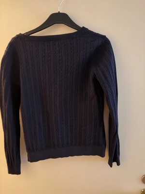 Mörkblå kofta från Bondelid, M - Snygg mörkblå kofta från Bondelid med kabelstickat mönster och v-ringning. Koftan har långa ärmar, knappar framtill och broderad logga med sköld på bröstet. Perfekt för dig som gillar klassisk och stilren look.