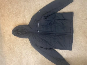 Svart pufferjacka Peak Performance - Svart pufferjacka från Peak Performance med huva och hel dragkedja framtill. Jackan har diskret quiltad design och logotyp på bröstet. Perfekt för kyliga dagar, med långärmade ärmar och snygg, enkel look.