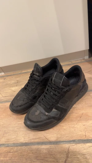 Valentino Garavani Rockrunner svarta sneakers - Svarta sneakers från Valentino Garavani med coolt camouflage-mönster och snygga detaljer. Skorna har snörning, platt sula och rund tå. Materialet är en mix av skinn och syntet, vilket ger en lyxig känsla och street vibe.