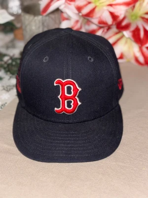 Boston Red Sox svart keps New Era - Svart keps från New Era med Boston Red Sox B-logga i rött och vitt framtill. På sidan finns en cool röd broderad grafik. Klassisk form med böjd skärm och ventilerande hål upptill. Perfekt för dig som gillar sportig streetstyle.