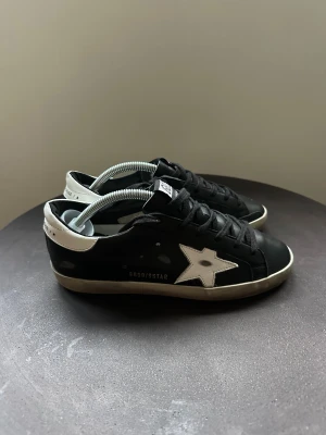 Svarta Golden Goose sneakers med stjärna - Snygga svarta sneakers från Golden Goose med vit stjärndetalj på sidan och vit häl. Skorna har snörning, rund tå och platt sula med mönstrad undersida. Materialet är skinn och detaljerna ger en cool och urban vibe. Perfekt för dig som gillar streetstyle.