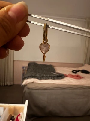Maria örhänge - Unikt örhänge i guldton med ett hjärtformat hänge i ljusrosa sten och en liten spetsig detalj nedtill. Perfekt accessoar för dig som vill sticka ut med något sött och trendigt.