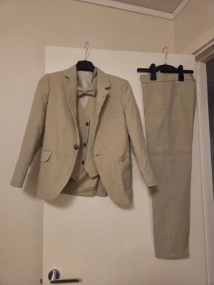 Beige kostym med väst och fluga för barn - Stilren beige kostym med kavaj, matchande väst och byxor samt en snygg fluga. Kavajen har klassisk passform, två knappar och fickor med lock. Västen har tre knappar och två fickor. Materialet är lätt och har en diskret struktur, perfekt för ljusa tillfällen.