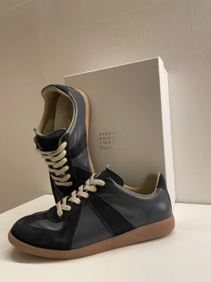 Maison Margiela mörkblå sneakers - Maison Margiela sneakers i mörkblå med beige snörning och gummisula. Ovandelen är en mix av skinn och mocka med snygga paneler och rund tå. Insidan är ljusbeige och yttersulan har ett unikt mönster med logotyp. Perfekt för dig som gillar stilrena och exklusiva sneakers. Priset är inte hugget i Sten!!!
