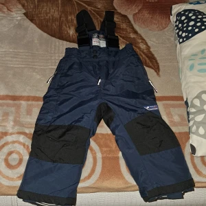 Mörkblå täckbyxor med hängslen - Säljer ett par mörkblå täckbyxor från Termo med svarta förstärkningar på knän och bak. Byxorna har justerbara svarta hängslen, dragkedja och tryckknappar framtill samt reflexdetaljer vid fickorna. Perfekt för kalla dagar och vinteraktiviteter.