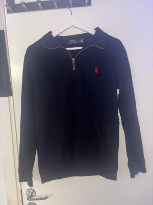 Svart quarter zip tröja Polo Ralph Lauren - Svart långärmad tröja från Polo Ralph Lauren med half zip och klassisk röd broderad logga på bröstet. Tröjan är stickad i bomull och har en stilren krage. Perfekt för dig som gillar enkel och snygg design.
