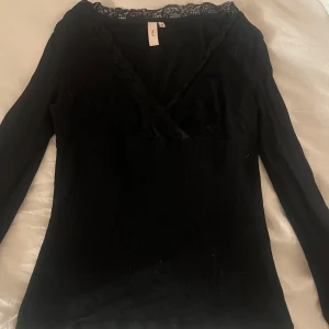 Svart omlottopp med spets  - Säljer en svart omlottopp från Nelly med v-ringning och fina spetsdetaljer längs halsen. Toppen har långa ärmar och är figurnära i modellen, perfekt för dig som gillar stilrena och feminina plagg.