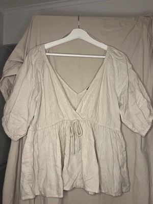 Beige omlottblus från Bik Bok - Säljer en beige omlottblus från Bik Bok i storlek M. Blusen har korta puffärmar, v-ringad hals och knytband i midjan. Tillverkad i ett lätt linnematerial som ger en chill och luftig vibe. Perfekt för dig som gillar en avslappnad men ändå snygg stil.