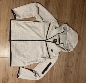 Grå vindjacka från Nike med huva - Grå vindjacka från Nike med svart logga på bröstet, hel dragkedja och huva. Jackan har svarta detaljer längs dragkedjan och ärmarna, samt en praktisk ficka med dragkedja på ena ärmen. Materialet är mjukt och känns sportigt, perfekt för chill eller träning.