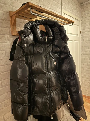 Moncler Montbeliard Hooded Short Down Jacket - Svart pufferjacka från Moncler med glansig finish, stor huva och klassisk logga på bröstet. Jackan har 2 olika dragkedjor framtill, fickor med lock. Letar du efter en Jacka som ska hålla dig varm, du har du hittat rätt🤩👌