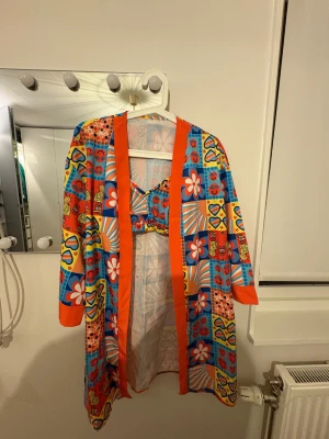 Färgglad bikini med retromönster - Säljer en supercool bikini med tillhörande kimono i färgglatt retroinspirerat mönster. Bikinin har mönster med hjärtan, blommor, smileys och solglasögon i orange, blått, gult och rött. Materialet är lätt och syntetiskt, perfekt för stranden. Kimono med orange kant och trekvartsärm ingår.