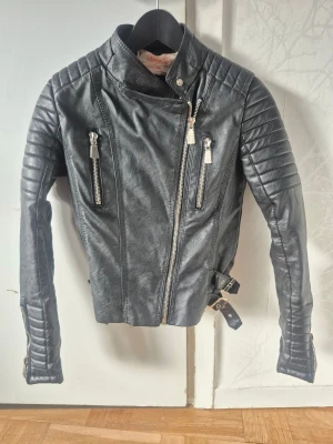 Svart skinnjacka från Voyelles, stl 36 - Snygg svart skinnjacka från Voyelles i bikerstil med quiltade detaljer på axlar och ärmar. Jackan har flera dragkedjor framtill, två spännen i midjan och ståkrage med tryckknapp. Perfekt för dig som gillar edgy och stilren look.