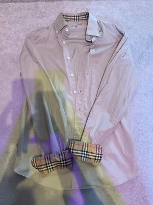 Beige Burberry skjorta - Snygg beige skjorta från Burberry i classic fit med ikoniskt rutigt mönster på insidan av krage och manschetter. Skjortan har knappar framtill, lång ärm och diskret Burberry-logga broderad på bröstet. Perfekt för dig som gillar stilrena och klassiska plagg. Typ nyskick och passar mig som har storlek M ungefär. Skriv om du har några frågor!