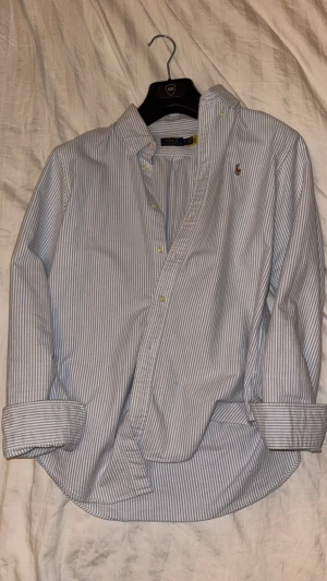  Polo Ralph Lauren skjorta - Klassisk skjorta från Polo Ralph Lauren med smala blå och vita ränder, broderad logga på bröstet och knappar framtill. Skjortan har lång ärm och är tillverkad i mjuk bomull. Perfekt för en stilren och avslappnad look. Storlek: M. Tar endast swish och köp sker utanför denna appen, dvs utan köparskydd!