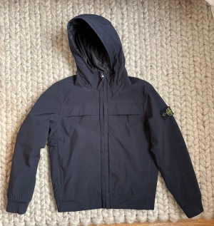 Marinblå fodrad vindjacka från Stone Island stl 10 år - Marinblå fodrad vindjacka från Stone Island Junior stl 10 år, med ikonisk patch på ärmen. Jackan har huva, dragkedja framtill och är tillverkad i slitstark polyester. Insidan är fodrad och jackan har en clean, stilren look som passar streetwear. Perfekt för dig som gillar en snygg och funktionell jacka. Inköpt på NK och inte särskilt mkt använd så skicket är så gott som nytt. Nypriset var 4200kr. 