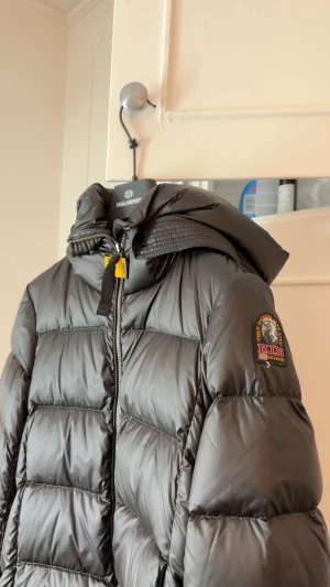 Svart lång dunjacka Parajumpers - Säljer en svart lång dunjacka från Parajumpers med huva och dragkedja. Jackan har klassisk quiltad design, patch på ärmen och snygga detaljer som gul rem vid halsen. Perfekt för kalla dagar och riktigt stilren look. Kvitto finns alla paper äkta!