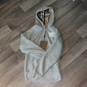 Grå hoodie från BB med dragkedja - Snygg grå hoodie från BB med klassiskt rutigt foder i huvan och praktisk dragkedja framtill. Tröjan har långa ärmar, ribbade muddar och snörning i huvan. Perfekt för dig som gillar stilren design med en touch av lyx. Sänker pris vid snabb affär och snabb frakt 