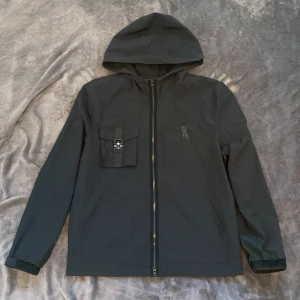 Svart vindjacka från Risky - Svart vindjacka från Represent med huva och hel dragkedja framtill. Jackan har en cool ficka med spänne på bröstet och ett diskret R-tryck. Tillverkad i lätt polyester, perfekt för blåsiga dagar. Elastiska muddar vid ärmslut och stilren design.