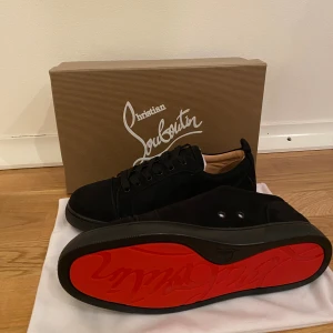 Svarta sneakers från Christian Louboutin - Säljer ett par svarta sneakers från Christian Louboutin med ikonisk röd sula. Skorna har rund tå, snörning och är tillverkade i mocka med svarta detaljer. Insidan är beige och Louboutin-loggan är broderad på hälen. Perfekt för dig som gillar lyxiga och stilrena sneakers.
