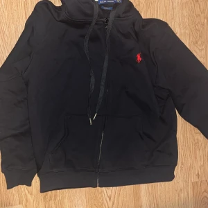 Svart hoodie från Polo Ralph Lauren - Svart hoodie från Polo Ralph Lauren med röd broderad logga på bröstet. Tröjan har huva med snörning, dragkedja framtill och två fickor. Det står storlek L på den fast det är M. Custom fit-modell som sitter snyggt och bekvämt. Luvan är också liten. 