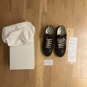 Maison Margiela GATS - Maison Margiela sneakers i svart med beige snörning och gummisula. Ovandelen är i skinn och mocka, med rund tå och platt sula. Klassisk och stilren design med diskreta detaljer på hälen. Levereras med originalkartong och dustbag.