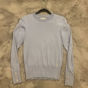 Ljusblå stickad tröja Holly & Whyte - Supersnygg ljusblå stickad tröja från Holly & Whyte med rund hals och långa ärmar. Tröjan har breda ärmslut med tre guldknappar. 