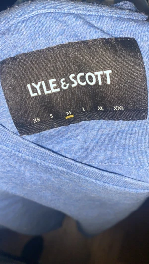 Blå t-shirt från Lyle & Scott, M - Snygg blå t-shirt från Lyle & Scott i storlek M, passar på S. Klassisk rund hals och korta ärmar. På bröstet sitter den ikoniska gula fågel-loggan. Tillverkad i mjuk bomull som känns skön mot huden. Perfekt för dig som gillar stilrena och enkla plagg.