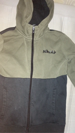 Nike Air hoodiejacka i olivgrönt och svart - Nike Air hoodiejacka med dragkedja, olivgrön upptill och svart nedtill. Jackan har huva, två sidofickor och mjukt fleecefoder på insidan. Nike Air-logga på bröstet. Perfekt för chill och streetwear.