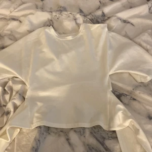 Vit croppad omlott-topp - Säljer en vit croppad topp med omlott-detalj baktill. Toppen har rund hals och korta ärmar, tillverkad i mjukt bomullsmaterial. Perfekt för dig som gillar minimalistisk stil och vill ha något enkelt men ändå med en cool twist.