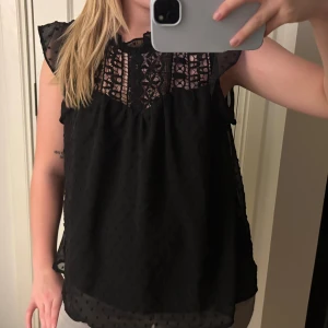 Svart blus med spets och volang - Svart blus med transparenta partier och spets upptill. Blusen har volangdetaljer vid axlarna och är ärmlös. Tyget är lätt och har små prickar i strukturen. Perfekt för dig som gillar en feminin och trendig look.