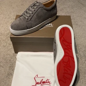 Gråa vita  Christian Louboutin skor - Snygga gråa sneakers från Christian Louboutin i mocka med klassisk röd sula. Skorna har rund tå, snörning och vit platt sula. Perfekta för dig som vill ha en stilren och lyxig look med en ikonisk touch.