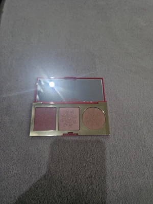 Estee Lauder Enchanted Glow palett - Superfin sminkpalett från Estee Lauder med tre produkter: en mörkrosa rouge, en skimrande highlighter och en ljus bronzer. Alla har snöflingeprägling och kommer i en röd och guldig ask med spegel. Perfekt för att skapa glow och färg i ansiktet.